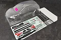An unpainted clear polycarbonate RC car body shell of a Skoda Fabia RS, including a decal sheet, rear wing, and window masks on a dark textured surface.