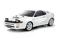 A studio shot of a white Tamiya Toyota Celica GT-Four ST185 1/10 scale RC car model with pop-up headlights and white wheels against a white background.