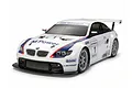 A Tamiya 1/10 scale RC BMW M3 GT2 race car model with white body, blue and red racing stripes, and sponsor decals on a white background.