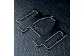 A black anodized aluminium battery and servo mounting plate for an RC car, featuring multiple screw holes and cutouts, resting on a dark textured surface.