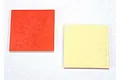 Two square pieces of double-sided mounting tape, one with a red backing and one with a yellow backing, on a white surface.