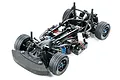 A high-angle studio shot of a Tamiya M-07 Concept 1/10 scale RC touring car chassis, showing the black plastic frame, suspension, and rubber tires.