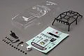 Axial 1/24 SCX24 Deadbolt clear unpainted body shell set including roll cage, decal sheet, driver helmet, and mounting hardware on a grey background.
