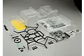 An unpainted clear polycarbonate RC crawler body shell kit with black plastic roll cage parts, light buckets, and decal sheets on a grey background.