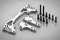 Rovan silver aluminium rear shock brace kit including three machined metal plates and a set of black steel screws on a grey background.