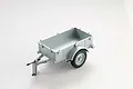 A 1/18 scale silver utility trailer for RC vehicles, featuring a single axle, rubber tires, and a hitch assembly with chains.