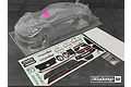 A clear, unpainted Bittydesign P-GT3R 1/10 scale RC car body shell with a decal sheet and small hardware bag on a dark surface.