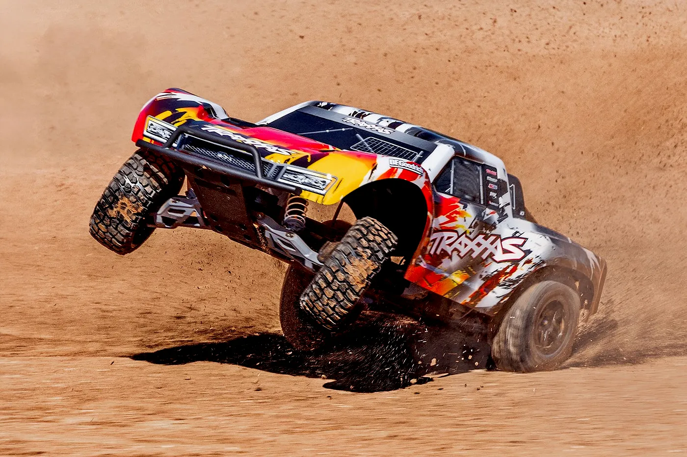 A Traxxas Slash short course RC truck performing a wheelie on a dirt track, kicking up dust.