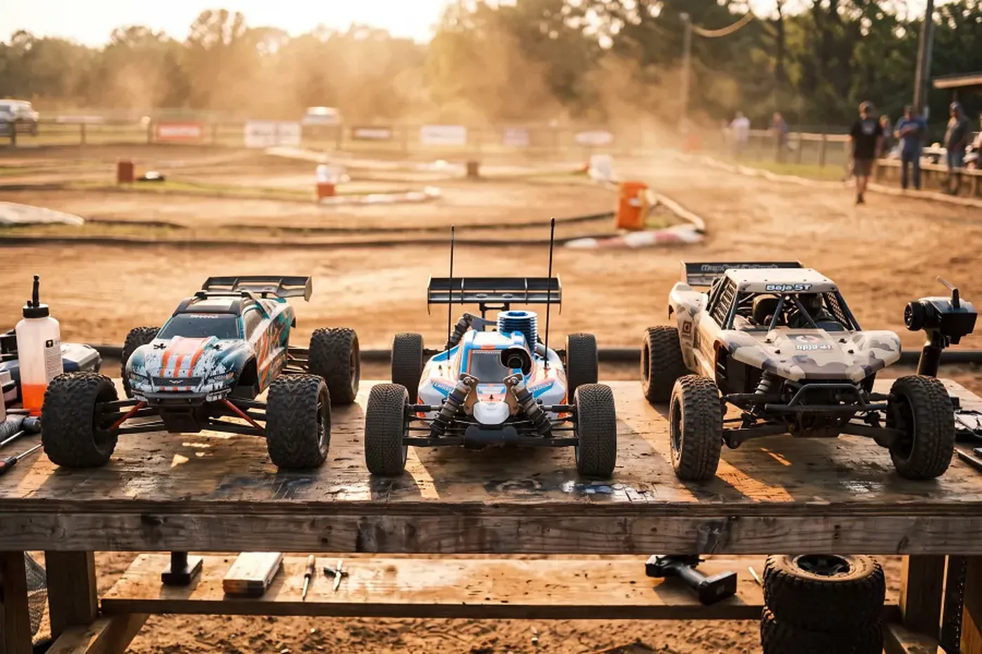 Three RC cars representing electric, nitro, and petrol power types lined up on a pit bench at an outdoor dirt track.