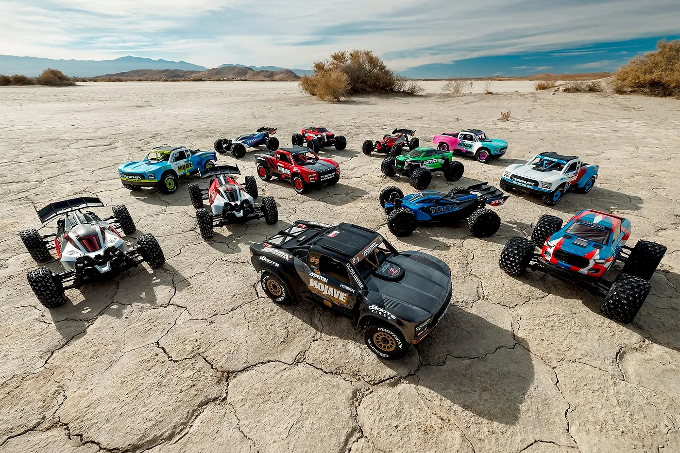 A large collection of various Arrma RC trucks and buggies arranged on a dry, cracked desert ground under a blue sky.