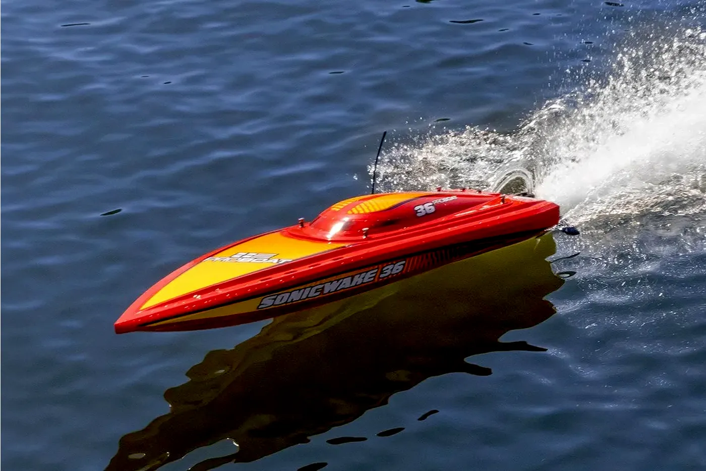 A red and yellow remote control speed boat racing across the water, kicking up a large spray of water from the stern.