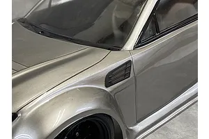 Close-up of a silver RC car body showing a black fender duct accessory installed on the front quarter panel.