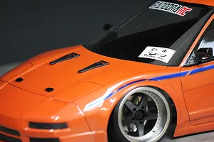 Close-up of an orange 1/10 scale RC car body featuring black NACA ducts installed on the hood.