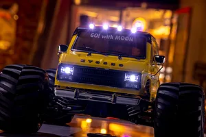 Front view of a yellow Kyosho Mad Van RC monster truck with Mooneyes branding, featuring illuminated headlights and roof lights in a dimly lit setting.