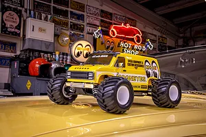 A yellow Kyosho Mad Van RC monster truck with Mooneyes branding sitting on the hood of a yellow car in a garage setting.