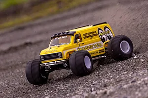 A yellow Kyosho Mad Van RC monster truck with Mooneyes branding driving fast on a gravel surface, kicking up dirt.