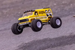A yellow Kyosho Mad Van RC monster truck with Mooneyes branding jumping in the air over a dirt surface.