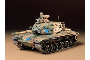 A fully assembled and painted 1/35 scale Tamiya M60A3 tank model with camouflage paint and a crew figure in the turret hatch.