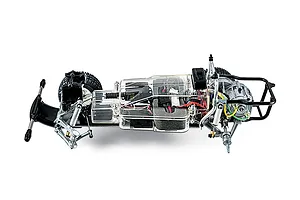 A side-profile view of a Tamiya 1/10 scale RC truck chassis kit, showing the internal components, clear plastic gearbox cover, and black metal frame.