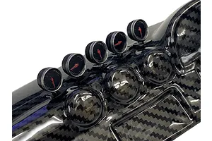Close-up of a carbon fiber textured RC dashboard interior featuring several small round analog gauges with red needles mounted on top.