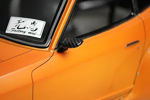 A close-up of a carbon fiber style side mirror mounted on an orange RC car body shell with a Factory mui sticker on the window.