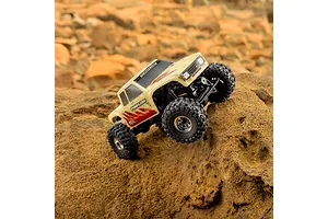 A tan topographical Pro-Line Coyote RC crawler body shell mounted on a chassis, climbing over a large brown rock outdoors.