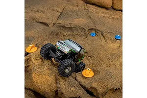 A remote-controlled rock crawler driving over a rocky terrain marked with blue and orange Pro-Line gate markers.