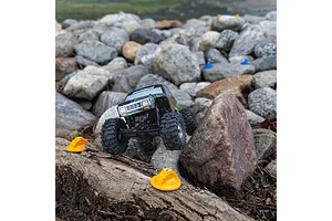 A small RC crawler vehicle driving over a log and rocks, with orange Pro-Line gate markers placed on the terrain.