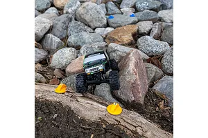A small RC crawler driving over a log with orange Pro-Line gate markers placed on the ground in a rocky outdoor terrain.