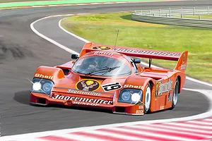 An illustration of a bright orange Porsche 956 race car with Jägermeister branding driving on a race track.