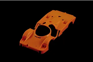 An unpainted orange plastic model car body shell for a Porsche 956, viewed from a high angle against a black background.