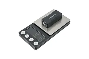 A black BetaFPV BT3.0 2S battery charger and voltage tester sitting on a digital weighing scale against a white background.