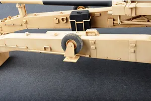 Close-up of the tan-colored chassis and support legs of a 1/16 scale M198 howitzer model kit, showing a small black storage box and a rubberized wheel.