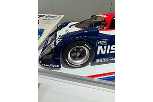 A close-up side view of a Tamiya Nissan R91CP RC car model on display, featuring blue, white, and red livery with Goodyear tire branding.