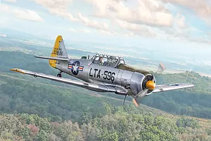 An artistic illustration of a silver T-6G Texan aircraft in flight over a forested landscape.