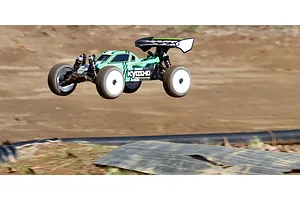 A green and black Kyosho Inferno MP10e RC buggy in mid-air during a jump on a dirt track.