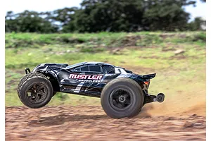 A black Traxxas Rustler RC stadium truck performing a wheelie on a dirt track outdoors.