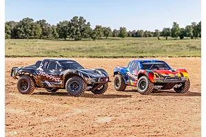 Two Traxxas Slash RC short course trucks, one black and one multi-coloured, parked on a dirt track outdoors.