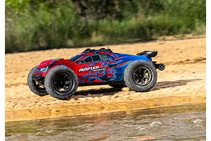A Traxxas Rustler 4X4 VXL RC stadium truck with a red and blue graphic body driving along a sandy riverbank.