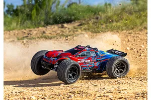A red and blue Traxxas Rustler 4X4 VXL RC stadium truck driving fast on a dirt track, kicking up dust.