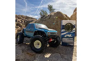 A blue RC crawler truck parked on a rocky outdoor trail next to a Pro-Line Rock-Tek tyre weight system product box.