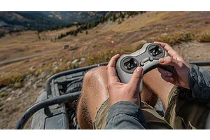 A person holding a grey CaddxFPV radio controller while sitting in an off-road vehicle with a mountain landscape in the background.