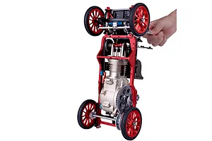 A hand holding a partially assembled Teching DM227 classic car model kit, showing the metal chassis, engine block, and red spoked wheels.