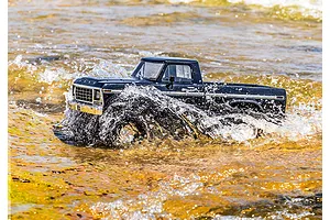 A black Traxxas TRX-4 1979 Ford F-150 RC truck driving through shallow, sunlit water, creating a large splash.