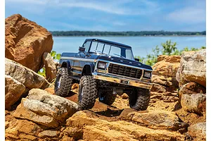 A black Traxxas TRX-4 1979 Ford F-150 Ranger XLT RC crawler driving over large orange rocks near a lake.