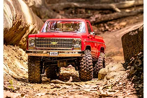 A red Traxxas TRX-4 1979 Chevrolet Cheyenne K10 RC crawler driving on a dirt trail between large rocks.
