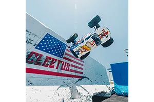 An ARRMA Freedom Flyer RC monster truck performing a high jump in front of a concrete barrier painted with an American flag and the word FLEETUS.