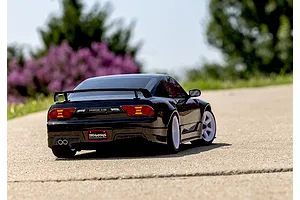 A black Traxxas Nissan 240SX RC car driving on a paved road with a blurred background of green trees and purple flowers.