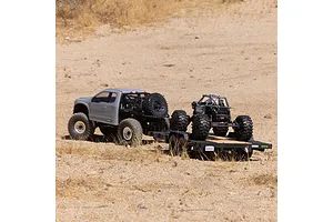 An Axial RC truck towing a flatbed trailer carrying a smaller RC rock crawler on a dry, sandy outdoor terrain.