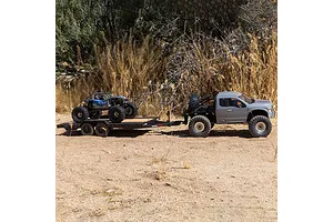 An RC flatbed trailer carrying a blue rock crawler, towed by a grey RC pickup truck on a dirt path.
